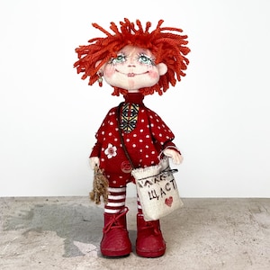 May include: A handmade doll with red yarn hair, wearing a red polka dot dress, striped socks, and red boots. The doll has a small bag with text and a heart. A small toy is held in the doll's hand. The doll has a smiling face.