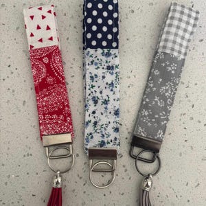 May include: Three fabric key fobs with metal hardware and tassels. One is red with white triangles and paisley, another is blue with white polka dots and floral print, and the third is gray with gingham and floral patterns.