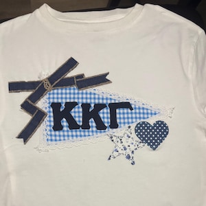 Puede incluir: Camiseta blanca con un diseño decorativo que presenta un banderín a cuadros azul y blanco con las letras "KKΓ", un lazo azul marino, una estrella y un corazón. El diseño está adornado con encaje.