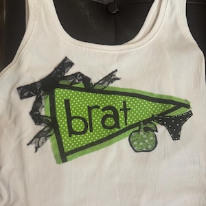 Puede incluir: Camiseta sin mangas blanca acanalada con un diseño de banderín verde y blanco con lunares. El banderín tiene la palabra "brat" en letras negras, un lazo negro y un gráfico de manzana verde.
