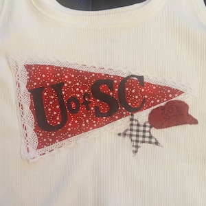 May include: White ribbed vest top with a red pennant displaying the letters "U of SC" in dark brown. The pennant has a white lace trim and a star pattern. A small red hat and a black and white star are also present.