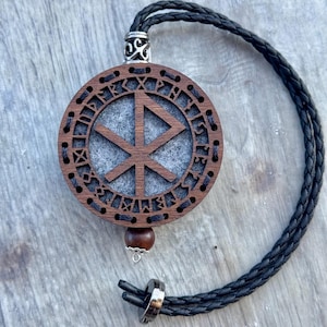 Könnte beinhalten: Runder Holzanhänger mit geschnitztem Design und grauem Mittelteil. Der Anhänger ist an einer schwarzen geflochtenen Kordel befestigt. Eine kleine Holzperle und ein silberfarbenes Metallstück befinden sich am unteren Ende des Anhängers.