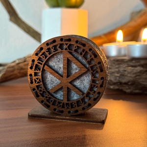 Pode incluir: Um porta-copos de madeira com um design circular esculpido com um símbolo central e runas externas. O porta-copos é castanho com um centro de feltro cinzento e assenta numa pequena base retangular. O design é provavelmente inspirado em temas nórdicos ou vikings.