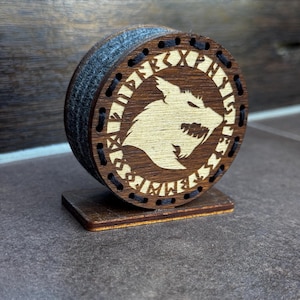 Könnte beinhalten: Ein runder Untersetzer aus Holz mit dunkelbraunem Finish, der in der Mitte eine lasergravierte Wolfskopf-Silhouette zeigt, umgeben von einem Ring aus Runenzeichen. Der Rand ist mit schwarzem Faden vernäht und steht auf einem rechteckigen Holzsockel.