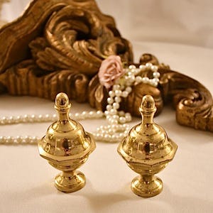 以下が含まれることがあります： 丸みを帯びたトップと溝の入ったベースを持つ、金色の金属製のフィニアルが2つ。フィニアルは白い表面の上に置かれています。