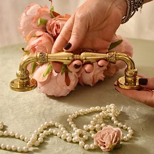 May include: A gold-toned door handle with a decorative design. The handle is shown against a background of pink flowers and a string of white pearls.