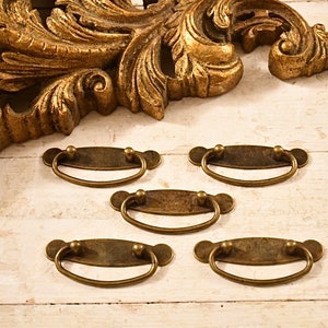 May include: Five antique brass drawer pulls with oval-shaped plates and rounded edges. The pulls have a distressed finish and are perfect for adding a vintage touch to furniture.