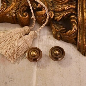 May include: Two gold-toned round cabinet knobs with a raised center and a decorative ring around the edge. The knobs are on a light beige surface.