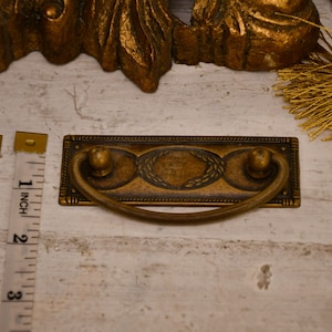 Puede incluir: Un tirador de cajón de latón con un diseño decorativo y una manija curva. El tirador mide aproximadamente 5 cm de largo.