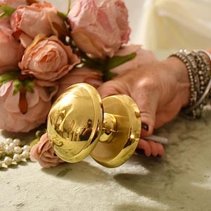 Puede incluir: Un pomo de puerta de oro pulido con un mango redondo y una base plana. El pomo se sostiene en una mano con flores rosas en el fondo.
