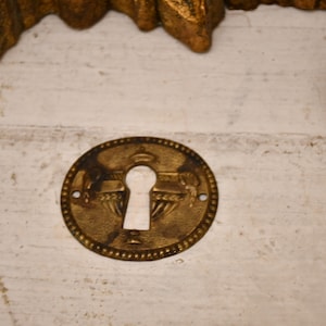 Op de afbeelding: Een ronde, messingkleurige sleutelgatbedekking met een decoratief ontwerp. Het sleutelgat bevindt zich in het midden van de afdekking en wordt omringd door een verhoogde rand.
