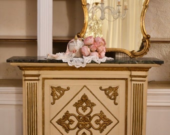 May include: A white and gold antique cabinet with a carved design on the front. The cabinet has a marble top and a mirror with a gold frame above it. A bouquet of pink roses is on the top of the cabinet.