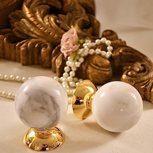 May include: Two white marble drawer pulls with gold bases. The pulls are round and have a smooth, polished finish.