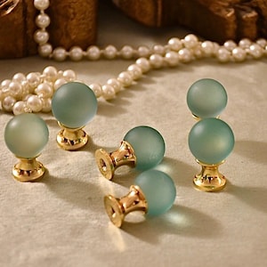 Puede incluir: Seis pomos de armario de vidrio esmerilado con bases de metal dorado. Los pomos son de color azul verdoso claro.