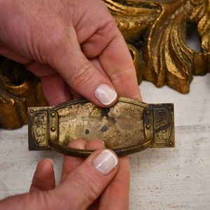 Puede incluir: Un tirador de cajón de latón con un diseño decorativo y un acabado ligeramente deslustrado. El tirador tiene una forma rectangular con una manija curva en el centro.