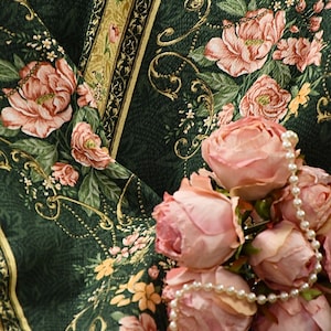 Op de afbeelding: Een witte parelketting gedrapeerd over een groene stof met een bloemenpatroon. De stof heeft roze rozen en gouden accenten.