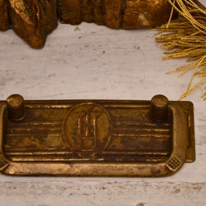 Puede incluir: Un tirador de cajón de latón con forma rectangular y dos pomos redondos. El tirador tiene un diseño decorativo en el centro, con un motivo estilizado.