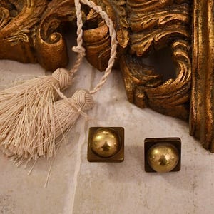 Puede incluir: Dos pomos de armario de metal dorado con un centro redondo ligeramente elevado. Los pomos son de forma cuadrada y tienen un aspecto ligeramente envejecido, antiguo.