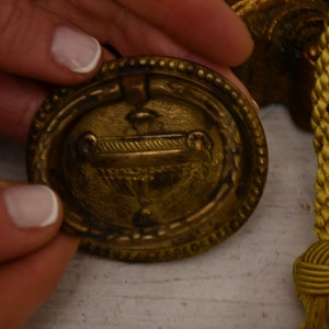 Puede incluir: Primer plano de un pomo de puerta de metal dorado con un diseño decorativo que presenta un cáliz o copa estilizada. El pomo tiene un borde con cuentas y un tirador de anillo.