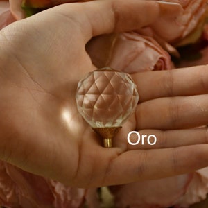 Puede incluir: Un pomo de cristal transparente con una base de metal dorado. El pomo está descansando en una mano, con la palabra "Oro" escrita debajo.