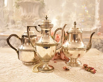May include: Three silver teapots with ornate designs and handles. The teapots are arranged on a white lace tablecloth with pink roses and a pearl necklace.