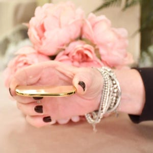 Puede incluir: Una mano sosteniendo un espejo compacto dorado con un ramo de flores rosas y blancas en el fondo.