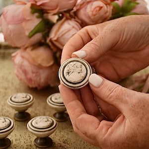 Peut inclure: Un ensemble de six boutons de cabinet en céramique blanche avec un motif en relief d'un visage de femme. Les boutons ont un anneau noir autour du bord et une base en bronze.