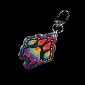Photograph of an acrylic keychain with a pride flag paw print design on a black surface lying flat on its back. Side covered in starry holographic coating is facing upwards and towards the camera.