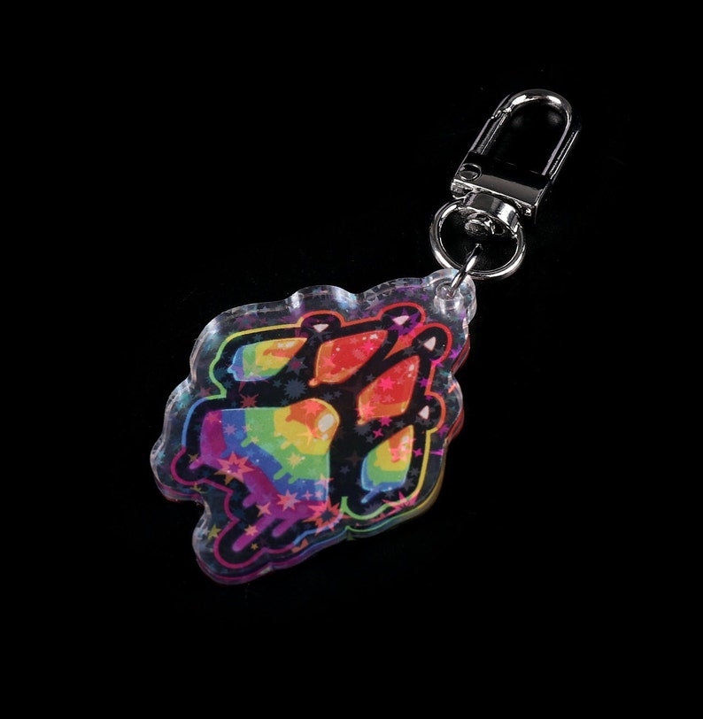 Photograph of an acrylic keychain with a pride flag paw print design on a black surface lying flat on its back. Side covered in starry holographic coating is facing upwards and towards the camera.