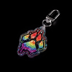 Photograph of an acrylic keychain with a pride flag paw print design on a black surface lying flat on its back. Side covered in starry holographic coating is facing upwards and towards the camera
