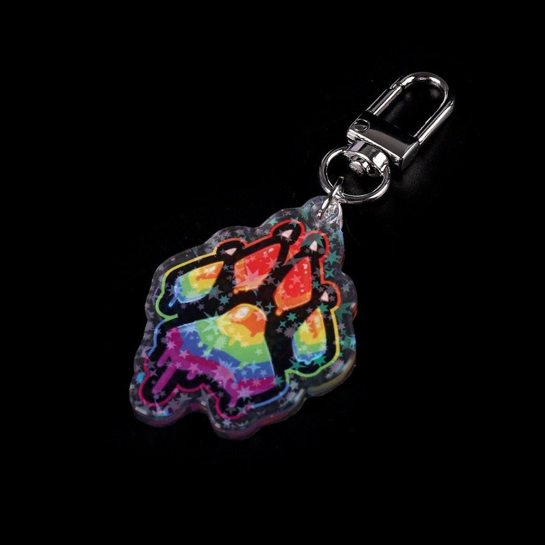 Photograph of an acrylic keychain with a pride flag paw print design on a black surface lying flat on its back. Side covered in starry holographic coating is facing upwards and towards the camera