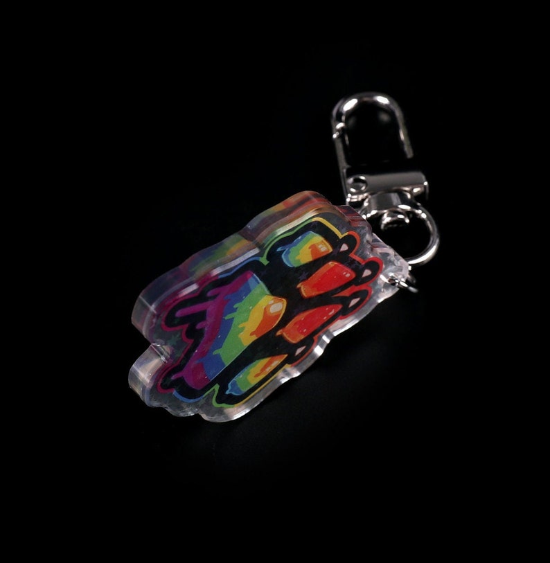 Photograph of an acrylic keychain with a pride flag paw print design on a black surface standing on it's side. Side not covered in starry holographic coating is facing forwards and towards the camera