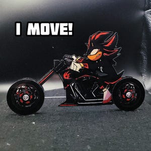 May include: A black and red character riding a black motorcycle. The character has red and black hair, an orange beak, and white gloves. The motorcycle has red accents on the wheels and frame. The text "I MOVE!" is in a white box above the character.