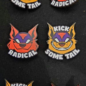 May include: A collection of colorful, cartoonish cat-like character pins. One pin features a red cat with the word "RADICAL" below. Another pin shows a yellow cat with the words "KICK SOME TAIL".