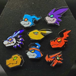 May include: A collection of nine colorful enamel pins featuring stylized character heads. The pins showcase a variety of designs, including a blue canine with an orange headband, a purple dragon, and a yellow rabbit. Each pin has a black outline.