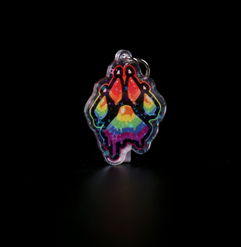 Photograph of an acrylic keychain with a pride flag paw print design on a black surface standing upwards. Side covered in starry holographic coating is facing forwards and towards the camera
