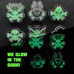 Może przedstawiać: Dziewięć świecących w ciemności naklejek z motywami potworów i skrzyżowanymi kośćmi. Naklejki są zielone i czarne, z zielonym napisem "WE GLOW IN THE DARK!". Naklejki mają różne twarze potworów.