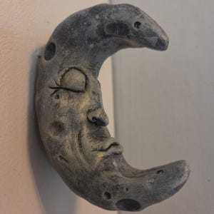 Pode incluir: Uma escultura de parede em forma de lua crescente com um acabamento cinza texturizado e desgastado. A lua apresenta um rosto humano esculpido com olhos fechados, nariz definido e lábios franzidos. A superfície tem pequenos cráteres.