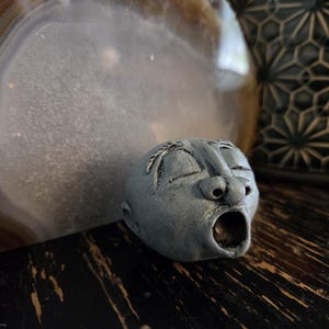 May include: A small, grey, sculpted ceramic head with closed eyes and an open, circular mouth. The sculpture features detailed facial contours, including a prominent nose and defined eyebrows, with a matte, textured finish on the surface.
