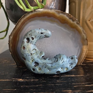 Op de afbeelding: Een blauwgroene halve maan sculptuur met een textuur en donkere gaten. De maan ligt op een grijsbruine agaat schijf. De sculptuur is een decoratief item.