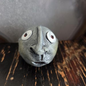 May include: A small, spherical gray sculpture of a face with a prominent nose, a small mouth, and two large, round white eyes featuring dark red pupils. The surface of the object has a textured, matte finish with visible contours and shading.