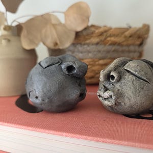 May include: Two small, rounded, hand-sculpted clay heads with textured, matte gray surfaces. Each head features stylized, abstract facial expressions with closed eyes, prominent noses, and defined lips, resting on a flat, coral-colored surface.