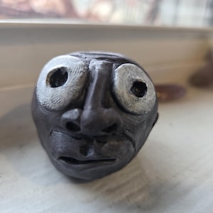 Könnte beinhalten: Eine dunkelgraue Keramikskulptur eines Gesichts mit großen, runden Augen und leicht geöffnetem Mund. Die Augen haben silberfarbene Ränder. Die Skulptur steht auf einer weißen Oberfläche.