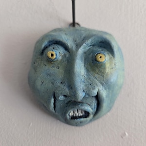 May include: A blue, sculpted face with yellow eyes and an open mouth revealing teeth. The face is mounted on a wall with a metal hook. The sculpture has a distressed, textured appearance.