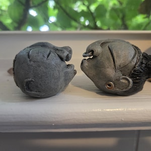 May include: Two small, gray, sculpted heads made of a matte material. The head on the left is rounded with carved features. The head on the right features a dark, textured hairstyle, a small metal ring in the nose, and a metal earring in the earlobe.