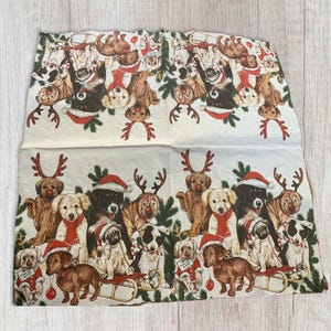 Könnte beinhalten: Eine weiße Papierserviette mit einem festlichen Weihnachtsdesign, das verschiedene Hunde zeigt, die Weihnachtsmützen und Rentiergeweihe tragen. Die Hunde sind von Stechpalmen und anderem weihnachtlichen Grün umgeben.
