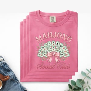 May include: Pink t-shirts stacked, featuring a graphic of Mahjong tiles arranged in a fan shape, with a pink bow and the words "MAHJONG Social Club" in a script font. The shirts are made of a soft material.