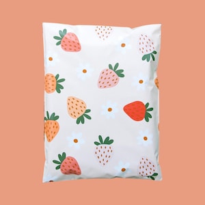 May include: A white poly mailer bag with a repeating pattern of strawberries in shades of pink and orange, along with white daisies. The strawberries have green leaves. The background is a light beige color.