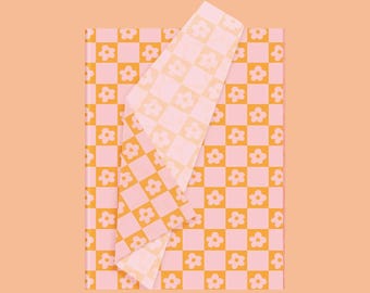 May include: A rectangular fabric piece featuring a repeating checkerboard pattern in light pink and orange. Each square contains a simple white five-petaled flower outline. The fabric is folded over at the top right corner, revealing the same pattern.