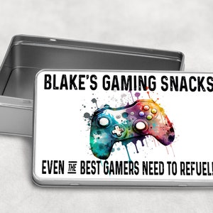 May include: A silver metal tin with a white lid featuring a colourful video game controller graphic and the text "BLAKE'S GAMING SNACKS EVEN THE BEST GAMERS NEED TO REFUEL!!"